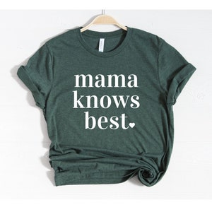 May include: A dark green t-shirt with the phrase "mama knows best" printed in white, with a small heart at the end. The shirt is hanging on a wooden hanger against a white background.