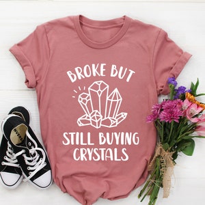 May include: A pink t-shirt with the text "Broke but still buying crystals" in white lettering. The text is surrounded by a graphic of white crystals.