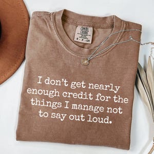 Puede incluir: Una camiseta marrón Comfort Colors con el texto "I don't get nearly enough credit for the things I manage not to say out loud." Un collar plateado con un colgante está sobre la camiseta.