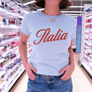 以下が含まれることがあります： 前面に赤い筆記体で「イタリア」とプリントされた薄い青色のTシャツです。