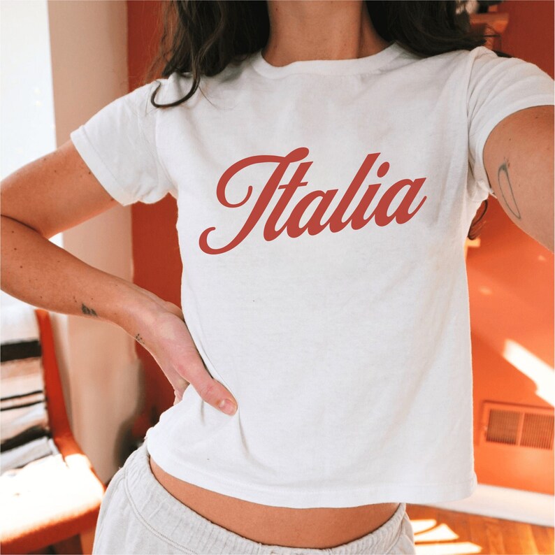 以下が含まれることがあります： 赤いイタリック体の「イタリア」のグラフィックが描かれた白いTシャツ