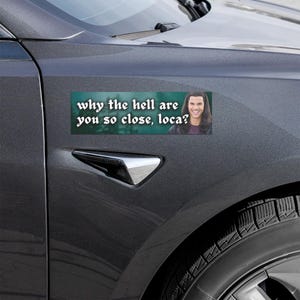 May include: A rectangular bumper sticker with the text "why the hell are you so close, loca?" in white font. The sticker features a photo of a smiling person. The sticker is attached to a gray car.