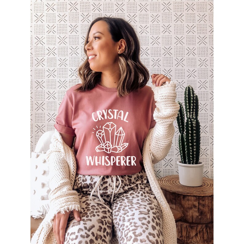 May include: A woman wearing a mauve t-shirt with the text "CRYSTAL WHISPERER" and a graphic of crystals. She is wearing leopard print pants and a white cardigan.
