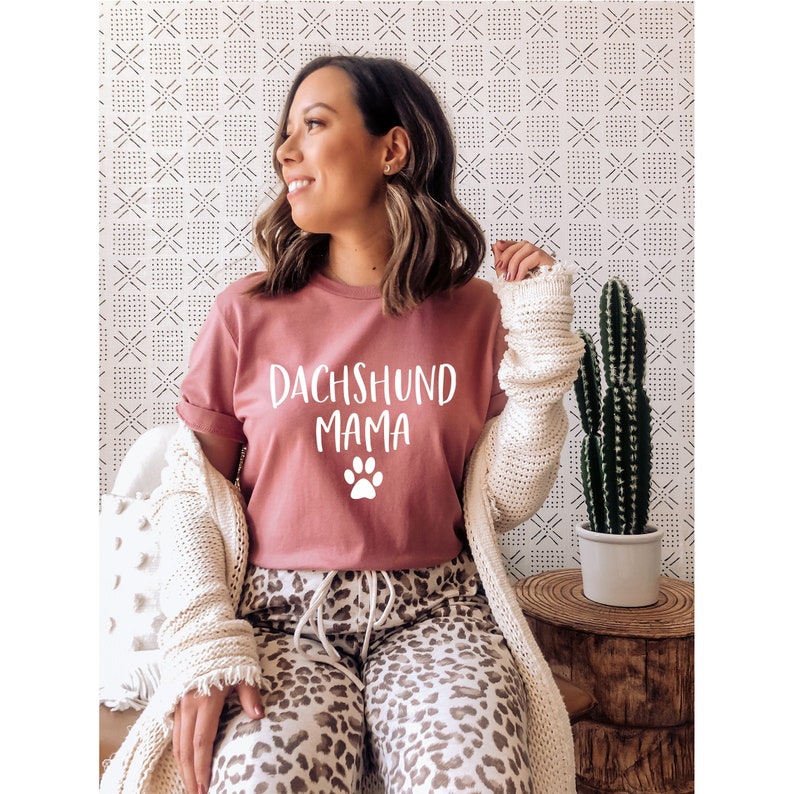 Op de afbeelding: Een vrouw draagt een roze T-shirt met de tekst "Dachshund Mama" en een witte pootprint. Ze draagt een wit vest en een broek met luipaardprint.