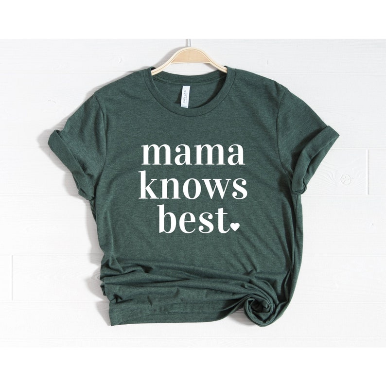 May include: A dark green t-shirt with the phrase "mama knows best" printed in white, with a small heart at the end. The shirt is hanging on a wooden hanger against a white background.