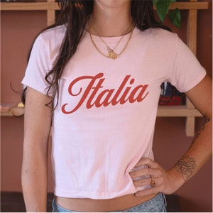 以下が含まれることがあります： 赤い筆記体で「イタリア」と書かれたピンクのクロップドTシャツです。