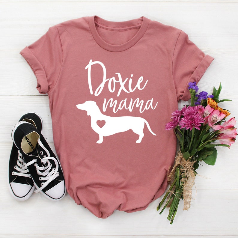 Puede incluir: Camiseta rosa polvoriento con las palabras "Doxie mama" y una silueta blanca de perro salchicha con un coraz&oacute;n. Zapatillas de lona negras y un ramo de flores junto a la camiseta.
