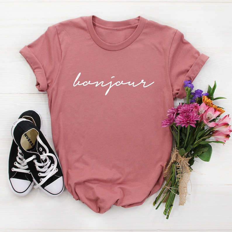 Pu&ograve; includere: Una maglietta rosa antico con la parola "bonjour" in corsivo bianco. La maglietta &egrave; abbinata a scarpe da ginnastica di tela nere e a un bouquet di fiori colorati, tra cui fiori rosa, viola e arancioni.