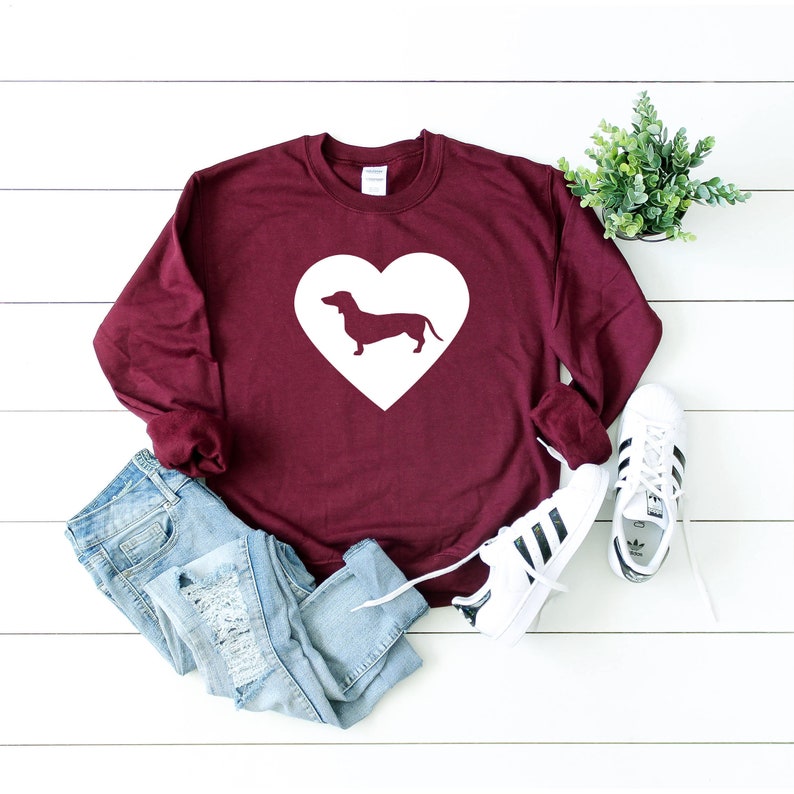 Op de afbeelding: Een bordeauxrood sweatshirt met een wit hartontwerp met een teckel-silhouet. Het sweatshirt wordt gecombineerd met een blauwe jeans en witte sneakers.