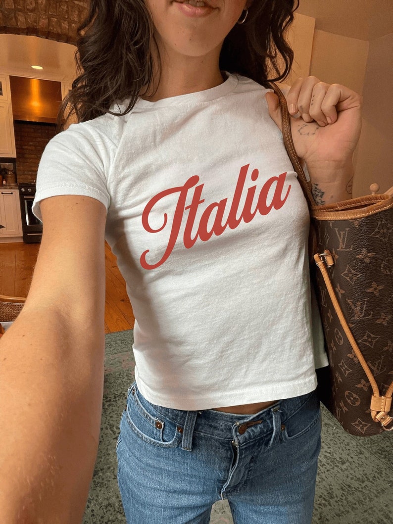 以下が含まれることがあります： 前面に赤い筆記体で「イタリア」と書かれた白いTシャツ。長い黒髪と青いジーンズを履いた人が着ています。