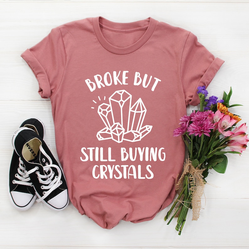 May include: A pink t-shirt with the text "Broke but still buying crystals" in white lettering. The text is surrounded by a graphic of white crystals.