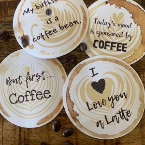 May include: Four white coffee coasters with coffee-themed designs and text. One says "My birthstone is a coffee bean." Another says "Today's mood is sponsored by COFFEE." The third says "But first... Coffee." The last says "I Love you a Latte."