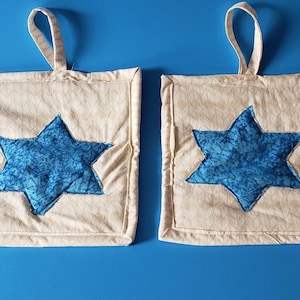 Puede incluir: Dos agarraderas de olla beige con diseños de estrella de David azules. Las agarraderas tienen lazos para colgar.