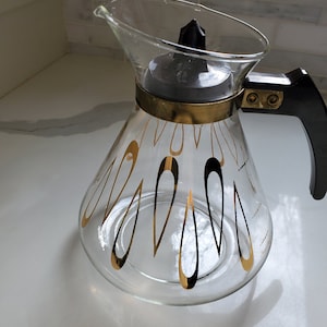 Puede incluir: Una cafetera de vidrio con un diseño dorado y negro. La cafetera tiene un asa de plástico negro y una banda de metal dorado alrededor de la parte superior.