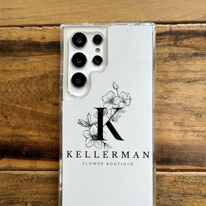 May include: A white phone case with a clear back. The case features a floral design with a black letter "K" in the centre. The text "KELLERMAN FLOWER BOUTIQUE" is printed below the design.