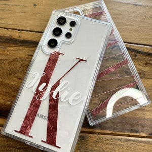 May include: A clear phone case with a white background and a large, red, glittery letter "K" with the name "Kylie" written in white cursive above it. The case is designed for a Samsung Galaxy phone.