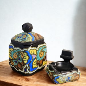 May include: A black ceramic jar with a lid and a small ashtray, both featuring colorful floral designs in blue, yellow, and brown. The jar has a geometric shape and a dark knob on the lid. The ashtray has a similar design.