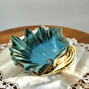 Peut inclure: Un bol en céramique en forme de coquillage, avec un intérieur turquoise brillant et un extérieur texturé, vert foncé et marron. Le bol est posé sur un tissu blanc bordé de dentelle, mettant en valeur son design et sa palette de couleurs uniques.