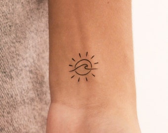 May include: A small, minimalist tattoo featuring a sun and wave design. The sun has radiating lines, and the wave is depicted with a single curved line. The tattoo is on the wrist.