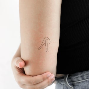 Op de afbeelding: Een eenvoudige lijntekening tattoo van een paar benen, met gebogen knieën, op de arm van een persoon.