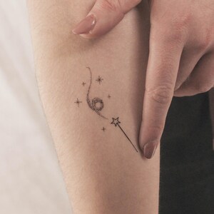 May include: A small, delicate tattoo of a swirling star with a wand and stars on a person's arm.