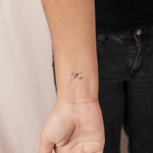 May include: A small, black tattoo of the cursive letter "K" on the wrist. The tattoo is delicate and minimalist, with fine lines. The skin around the tattoo is fair-toned, and the background is blurred.