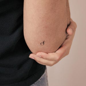 May include: A small, black, cursive letter S temporary tattoo applied to the skin. The design features a delicate, flowing script font with elegant curves, positioned clearly on the surface of the skin as a minimalist body art element.
