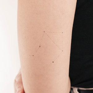 May include: A close-up of a person's arm with a small, delicate tattoo of the constellation Libra. The tattoo is done in a minimalist style with small dots and stars.