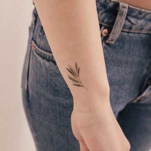 Puede incluir: Un pequeño tatuaje delicado de una rama de hoja verde en la muñeca de una persona.