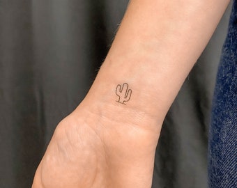 May include: A small, minimalist cactus tattoo on a wrist. The tattoo is a simple outline in black ink, depicting a stylized cactus with three arms and a base. The skin tone is fair, and the background is a dark, blurred fabric.