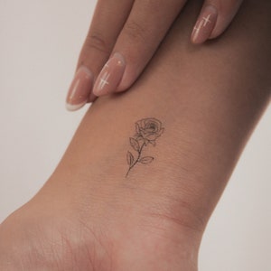 Puede incluir: Un simple dibujo de línea de un tatuaje de rosa en la muñeca de una persona.