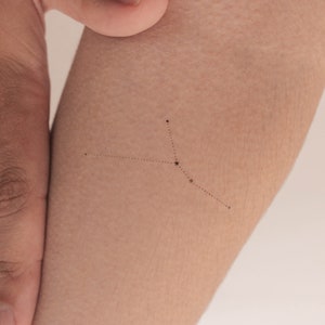 May include: A small, simple tattoo of the Cancer constellation on a person's arm. The constellation is made up of small dots connected by thin lines.