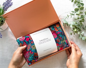 May include: An open peach-colored gift box reveals a floral patterned eye pillow. The pillow is teal with a vibrant pattern of pink, red, and orange flowers. A white label reads "holme&moss" and describes the product as a relaxing eye pillow.