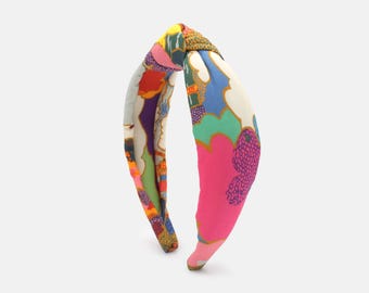 May include: A colorful headband with a knotted top. The fabric features a vibrant pattern with pink, blue, green, and orange hues. The headband is designed for hair styling and fashion.