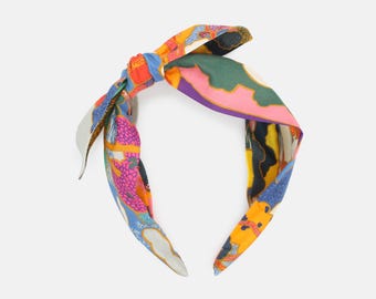 May include: A colorful headband with a knotted bow. The headband features a vibrant pattern with shades of blue, orange, pink, and green. The design includes abstract shapes and floral elements, creating a stylish accessory.