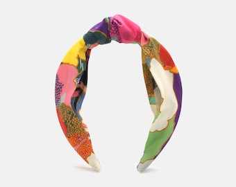 May include: A colorful headband with a knotted top. The fabric features a vibrant pattern of pink, yellow, green, purple, and orange shapes. The headband is designed for hair styling and fashion.