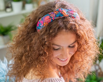 May include: A woman with curly brown hair is wearing a colorful floral headband with a knotted bow. The headband is made of fabric and has a variety of colors, including pink, blue, and green.