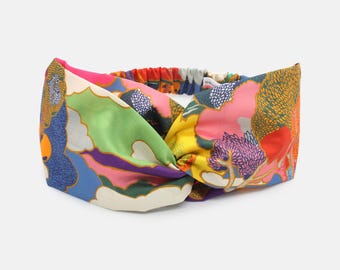 May include: A wide fabric headband featuring a twisted front design and an elasticized back strap. The surface displays a vibrant, multi-colored abstract pattern with shades of pink, yellow, green, blue, purple, and orange in various shapes.