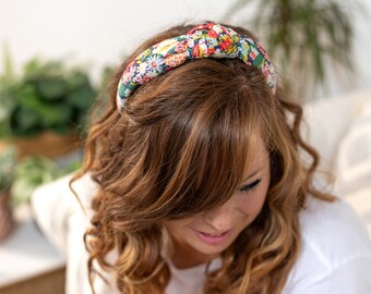 May include: A floral patterned headband with a knotted design. The headband is made of fabric and has a dark blue background with colorful flowers.