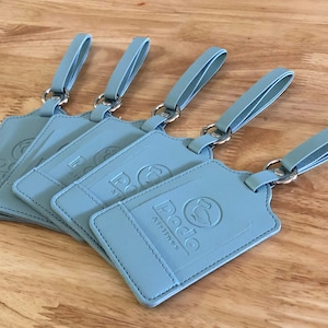 Puede incluir: Cinco etiquetas de equipaje de cuero azul claro con anillos y correas plateadas. Las etiquetas tienen el logotipo "Pedo Airlines" en relieve.