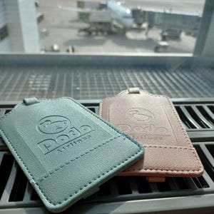 Puede incluir: Dos etiquetas de equipaje, una turquesa y otra marrón claro, con un logotipo de pájaro estilizado y el texto "Podo Airlines".