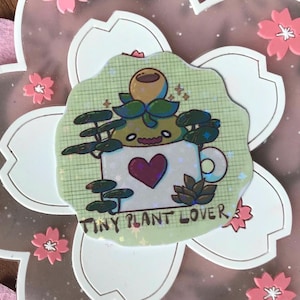 Könnte beinhalten: Ein holografischer Aufkleber mit einer Cartoon-Pflanze in einer Tasse mit einem Herzen. Der Aufkleber trägt den Text "TINY PLANT LOVER" und ist von dekorativen Elementen umgeben, darunter rosa Blüten.
