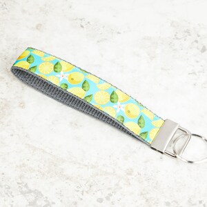 May include: A wristlet keychain with a vibrant lemon pattern on a light blue background. The fabric wrist strap features yellow lemons, green leaves, and white blossoms. The wristlet is attached to a silver-colored metal clasp and key ring.
