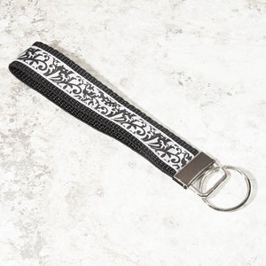 May include: Black wristlet key fob with a white and black floral pattern. The key fob has a silver metal clasp and a silver key ring. The wrist strap is made of a woven material.