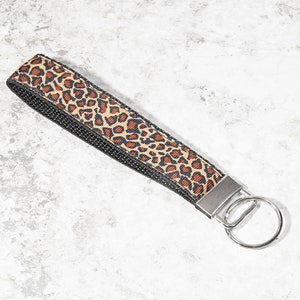 May include: A wrist key fob with a leopard print design in brown and black. The key fob has a black woven backing and a silver-colored metal ring for attaching keys.