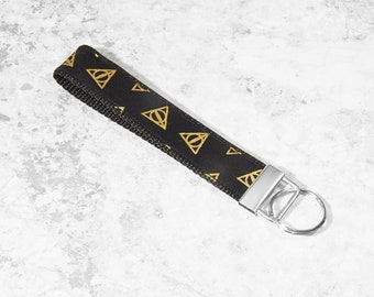 May include: Black wrist lanyard with gold Deathly Hallows symbols. The lanyard has a silver metal clasp and key ring. The fabric is a woven material. The design is a repeating pattern of the Deathly Hallows symbol.