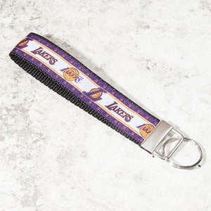 May include: A white and purple key chain with the Los Angeles Lakers logo and the word "LAKERS" repeated on the strap. The key chain has a silver metal ring.