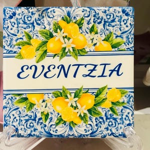 May include: Square ceramic tile with a blue and white decorative pattern. The tile features a lemon and floral design with the word "EVENTZIA" in blue script. The tile is displayed on a clear stand.