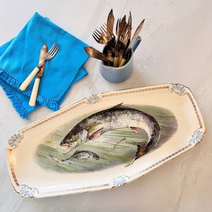 May include: A white ceramic serving platter with a gold trim and a detailed illustration of a fish. The platter has a decorative border with a repeating pattern.