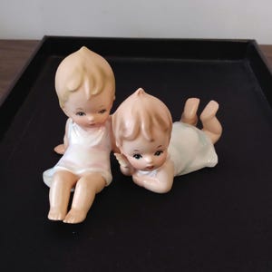 May include: Two porcelain figurines of babies. One is sitting, the other is lying down. Both have blonde hair, rosy cheeks, and are wearing white dresses. The figurines are on a black tray.